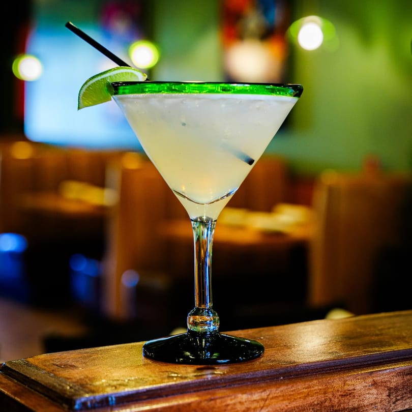 Margarita in a green-rimmed glass with a lime wedge and straw on a wooden bar.