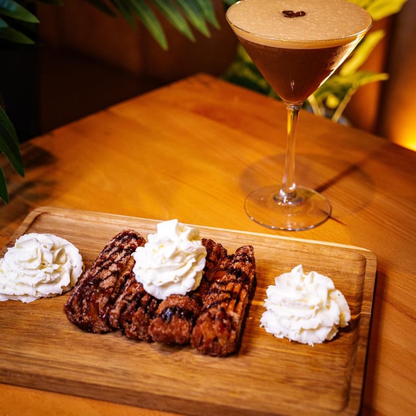 Churros with whipped cream and chocolate drizzle on a wooden board, with an espresso martini.