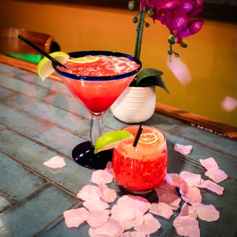 Two vibrant red cocktails, garnished with lime, on a blue tiled surface with pink petals.