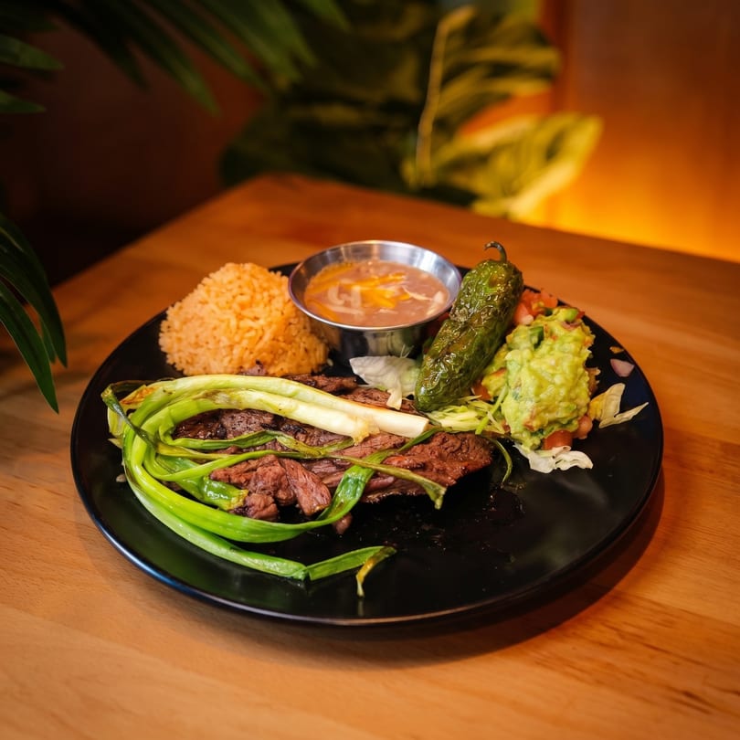 Grilled steak with green onions, rice, beans, guacamole, and a large green pepper on a black plate.