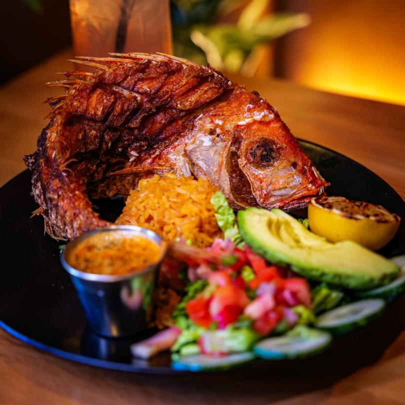Whole fried fish with rice, avocado, salad, and sauce on a black plate.