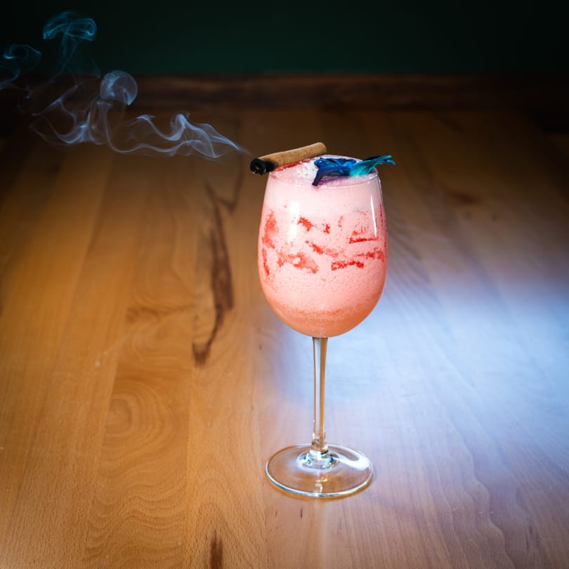Pink cocktail with a cinnamon stick and blue garnish, emitting smoke on a wooden table.