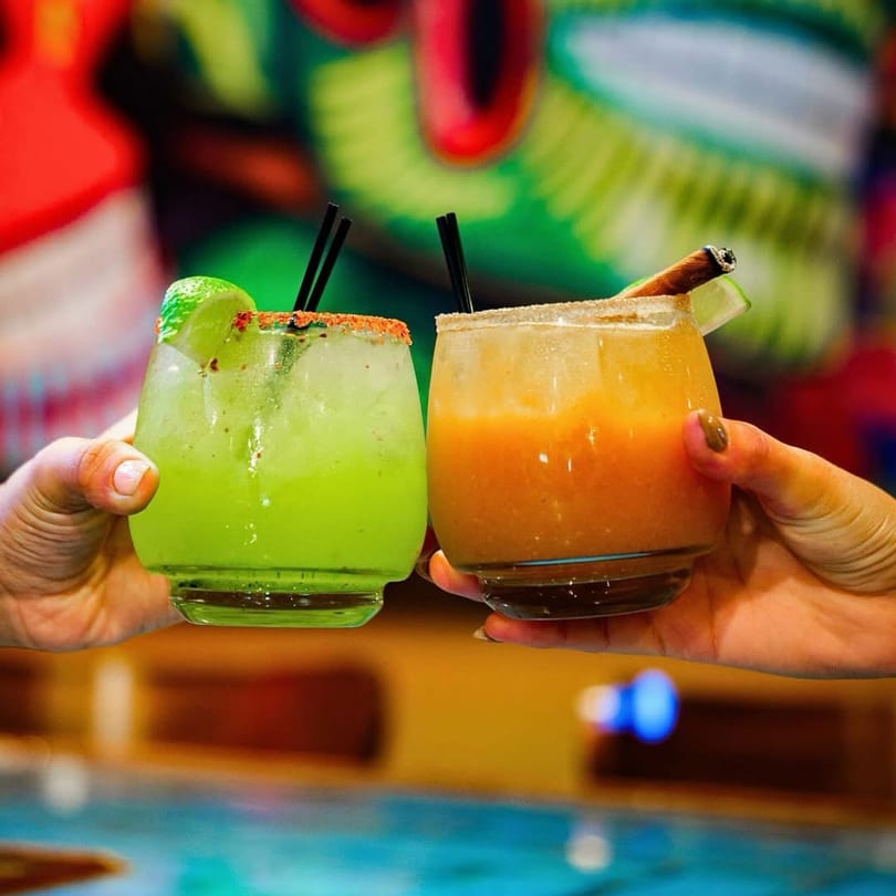 Two hands holding up vibrant green and orange cocktails with colorful garnishes against a blurred background.