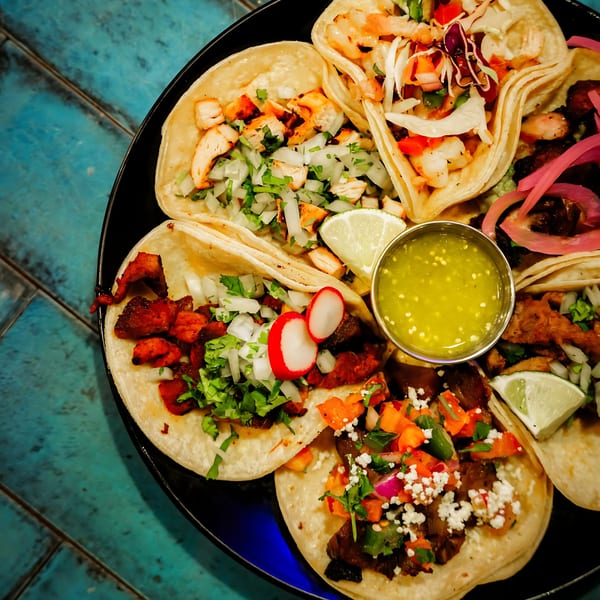 A black plate filled with various colorful tacos, a side of green salsa, and lime wedges.