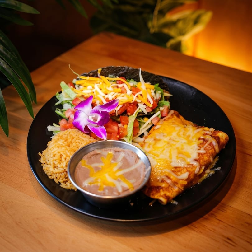 A black plate holds a taco, enchilada, rice, and refried beans, garnished with a purple orchid.