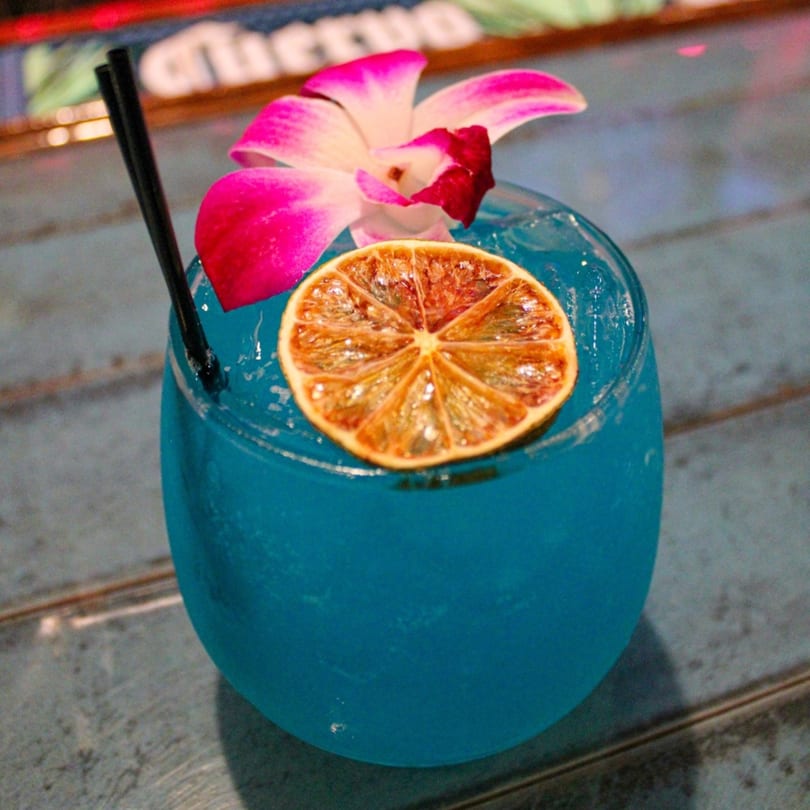 A vibrant blue cocktail garnished with a dried orange slice and a pink orchid.