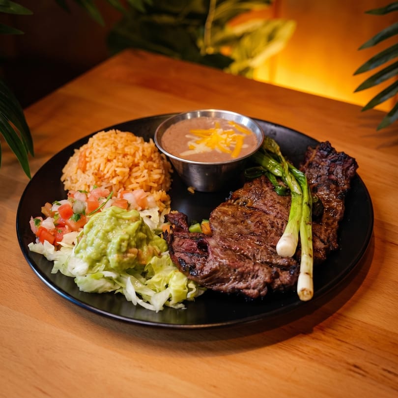 Grilled steak with rice, refried beans, guacamole, pico de gallo, and grilled green onions on a black plate.