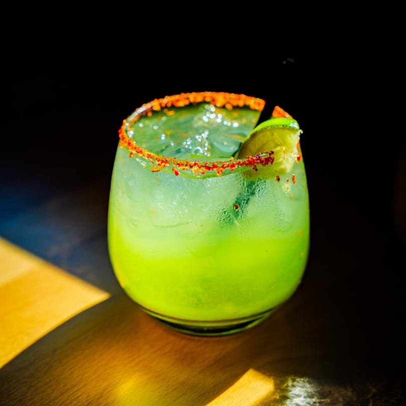 A vibrant green cocktail with a chili-salt rim and lime wedge, on a dark table with sunlight.